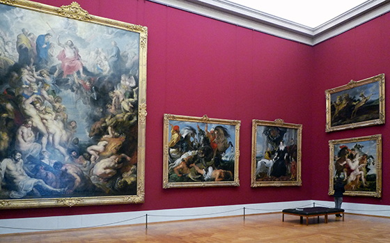 A photograph of 5 paintings, four of them approximately the height of the man who is standing in front of them in the photograph. The fifth is twice the size of the others. The paintings depict various scenes and topics.