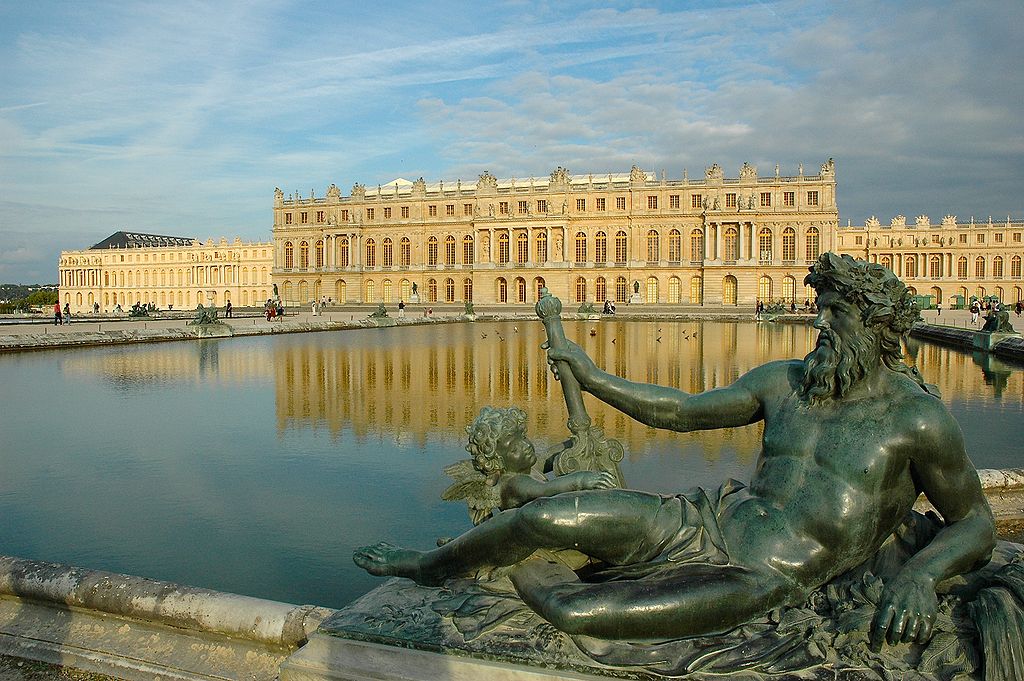 A statue lounges at the edge of a large artificial moat. On the far side of the water is the palace Versailles. The building is expansive, with two different songs visible from this side.