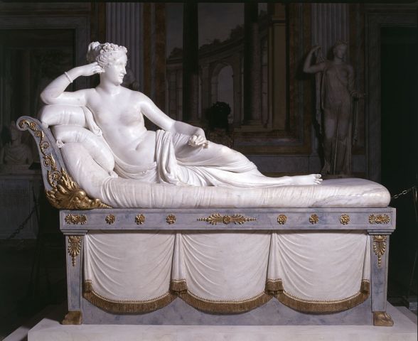 A sculpture. A woman half-nude, with a cloth covering the lower half of her body. She partially reclines on a bed with pillows supporting her. The bed is ornately carved of a blue-grey stone and gilded with gold.