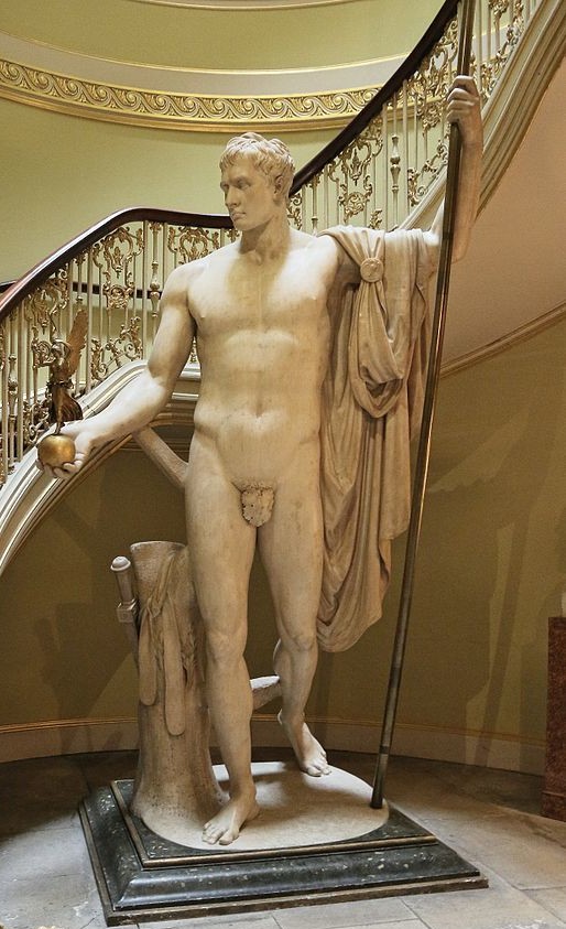 A nude man standing upright in a contrapposto position. He has an apple in his right hand. His left holds a spear aloft (the base of the spear is on the ground). He has a cloak draped over his arm.