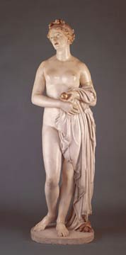 A nude woman standing upright, her arms folded in front of her. She holds a golden apple in her left hand. There is a swath of fabric folded over her left arm, draping to the ground, partially covering her lower body.