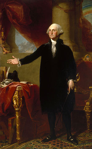 A full portrait of George Washington. He stands with his feet shoulder-width apart, with his right hand extended in an open gesture. His clothes are a plain black and white, but the room behind him is more richly garbed in reds and golds. The exposed leg of a table at George's side is exquisitely carved, with eagle details clearly prominent.
