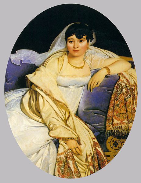 Una mujer relajada en una silla acolchada. El cojín es de un azul rico. La mujer lleva un vestido blanco con un chal color crema sobre el brazo.