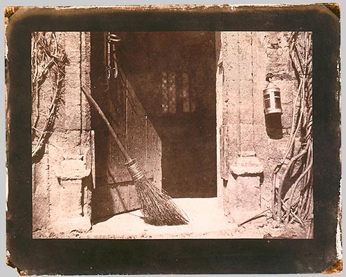 An open doorway with a broom leaning on one side of the frame. A lantern hangs by the other side of the door.