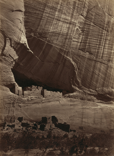 Ancient ruins in front of a vast canyon wall. The ruins are dwarfed by the scope of the wall's size.