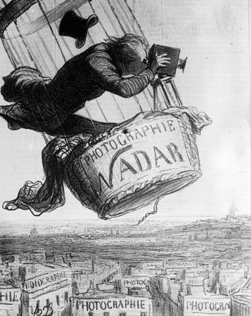 Nadar in a hot air balloon, taking photographs of the city below. His hat has flown off his head, and he looks precariously balanced.