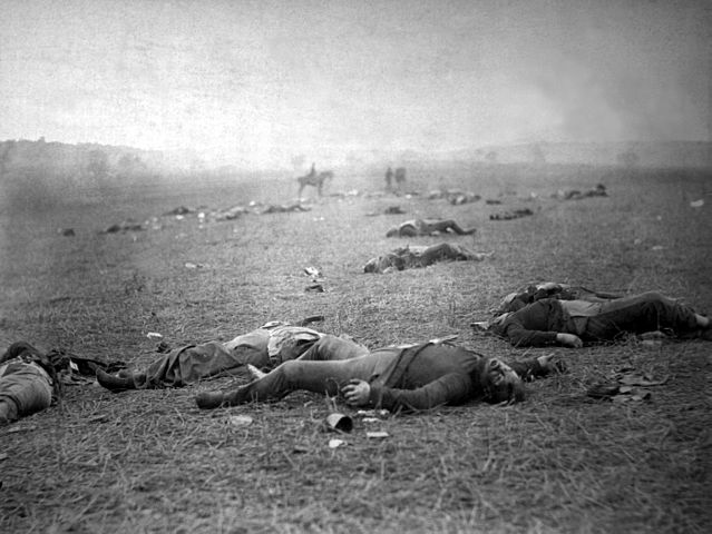 One soldier's body lies at the center of the photograph. A few dozen other men can be seen scattered across the ground. In the background of the photo, a man on horseback sits in the middle of the scene.