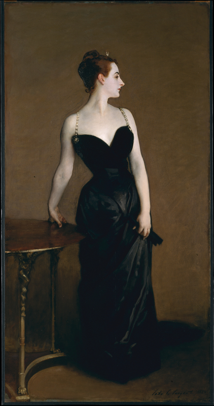 A full body portrait. A woman stands, wearing a floor length black dress with a sweetheart neckline and jeweled straps. She looks away from the viewer, with her face in profile.