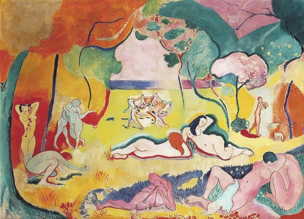 A scene of several nudes relaxing on a grassy field. The scenery around the people is abstract and the trees that line the field blend into one another. The color palette is bright, and not entirely realistic. Some individuals have typical Caucasian coloring, but others are painted in greenish hues, while others are almost purple. In the center of the background, several figures are holding hands and dancing in a circle.