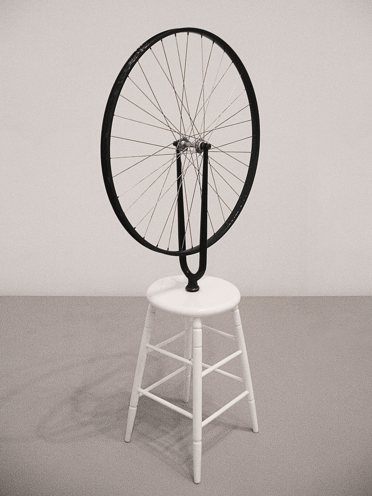 Un taburete blanco de cuatro patas con una rueda de bicicleta fijada al centro del asiento. La rueda está erguida.