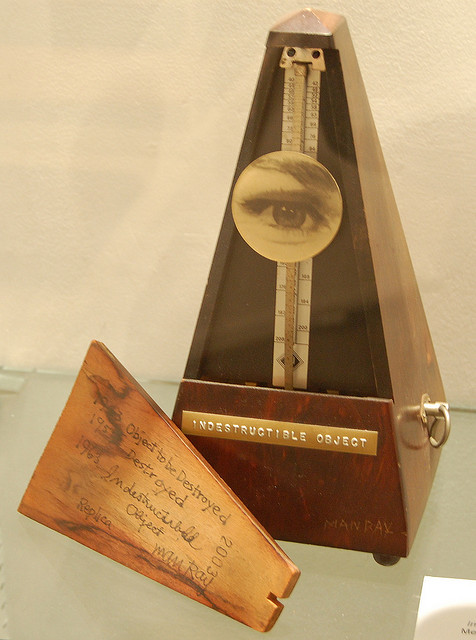 A metronome with a circular photograph of an eye and an eyebrow attached to its pendulum. The front of the metronome has a label stamped into metal: Indestructible Object.