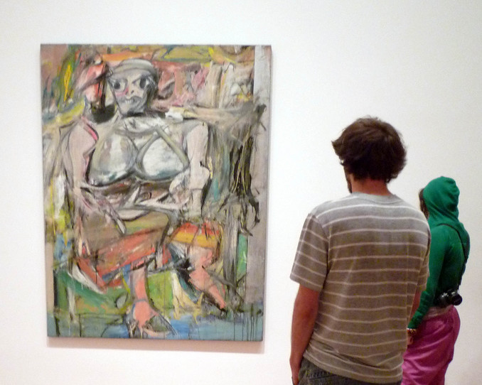 Un hombre y una mujer parados frente a Mujer I en exhibición en un museo. la obra de de Kooning muestra a una mujer sentada. La piel de la mujer es principalmente gris, y su cuerpo está simplificado y no anatómicamente proporcionado.