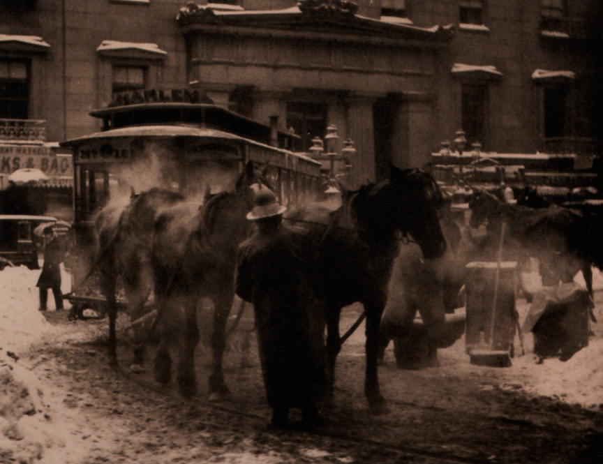This sepia photograph of a street features horse-drawn transportation as its focal point. A man stands in front of the two horses tending to them. The ground is covered in a thin layer of dirty snow, with drifts of cleaner snow piled along the sides of the road. Hot air is visible as mist coming from the horses’ mouths and bodies.