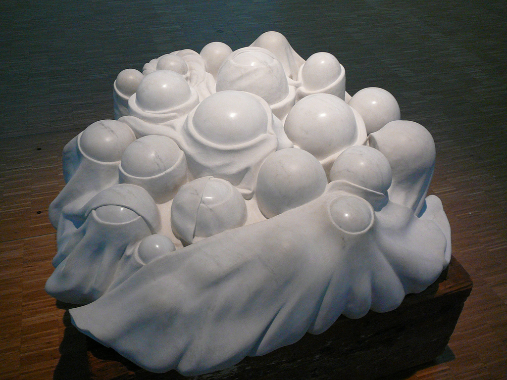 A marble sculpture seen at an angle from above. There are several spherical protrusions rising from a vaguely circular base. These protrusions are different sizes and rise to different heights, but are fairly uniformly round. It appears as though a sheet (also carved from the marble) has been draped over them, but the sheet has holes, exposing the tops of each sphere. There are several layers of the sheeting.