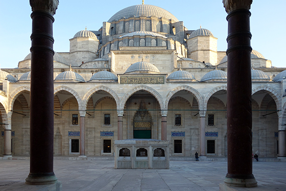 The photograph is framed with two columns in the foreground, illustrating the fact that the columns visible on the other side of the courtyard surround the entire interior. At the rear of the courtyard (the focus of the photograph) we see the main structure of the mosque. There is a central dome at the top, with two smaller immediately on either side of it. Below this is another dome shorter than the central dome, but approximately the same width; it is also framed by two smaller domes, though these are wider than the two above it. Just below this, there are several smaller domes covering the top of the courtyard’s edge.