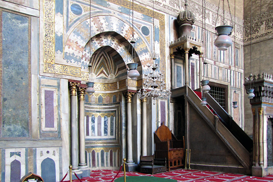 An intricate alcove with blue, gold, and bronze geometric patterns. Next to the alcove there is a wooden chair, and next to this there is a narrow staircase. The top of this staircase is sheltered by a roof with a large, intricate onion-shaped crown on its top. The roof is supported by columns, so the standing area is not obstructed.