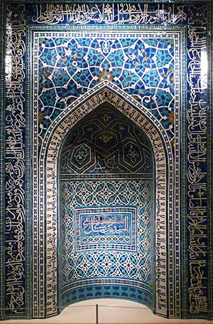 A prayer niche made up of blue, aquamarine, white, and gold tiles. The interior of the niche has arabic text and geometric patterns. The wall outside the niche has a rectangular frame outlining the arched niche entrance. Between the arch and the frame there is a geometric, almost floral pattern. Outside the frame there is more stylized arabic writing.
