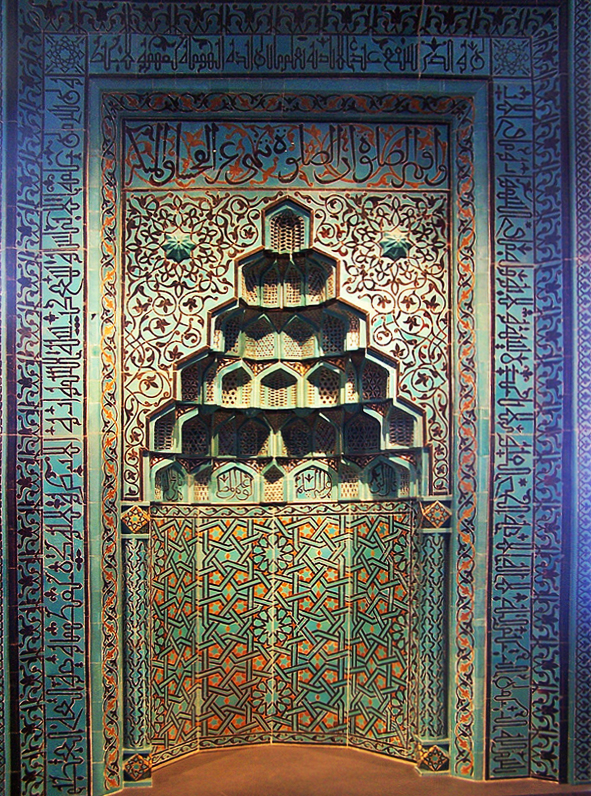 Intricate niche inset in a wall. The top niche is cut out in layers; It has an angled dome shape. The niche and surrounding walls are primary aquamarine with blue, maroon, and orange designs. These designs are geometric and include arabic writing.