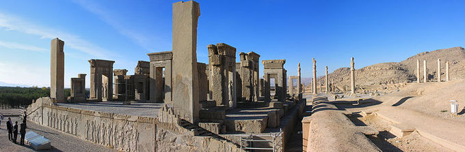 Photo depicts the ruins of Persepolis. It shows a raised foundation with steps on either side. On the foundation, there are various structures that look like doorframes.