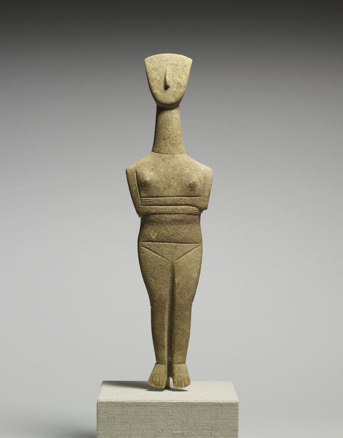 This is a color photograph of a Cycladic female figure, made of marble. It is from the Cyclades, Greece, circa 2500 BCE. The woman is sculpted with her legs together and arms folded over her abdomen, with her breasts and pubic region emphasized.
