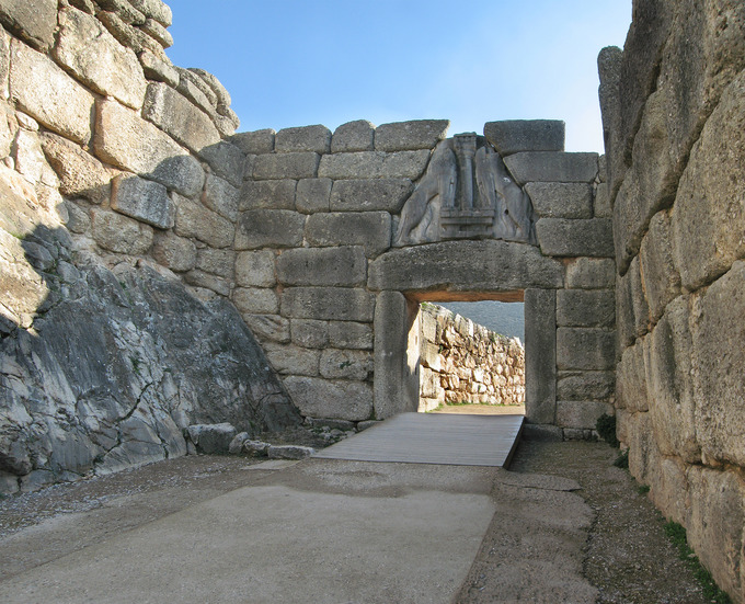 Color photo depicting a stone wall with an archway formed by two large stone pillars capped with a horizontal support. Atop the support, there's a relief of two lions facing one another.