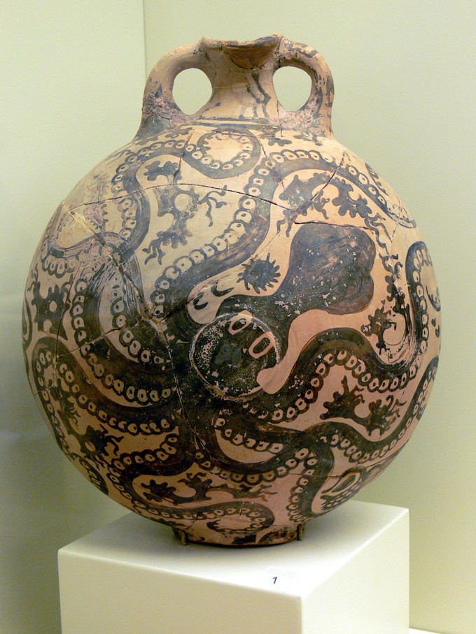 This is a color photograph of a pottery jug or vase. Its surface is covered by an octopus; bits of seaweed fill the negative space.