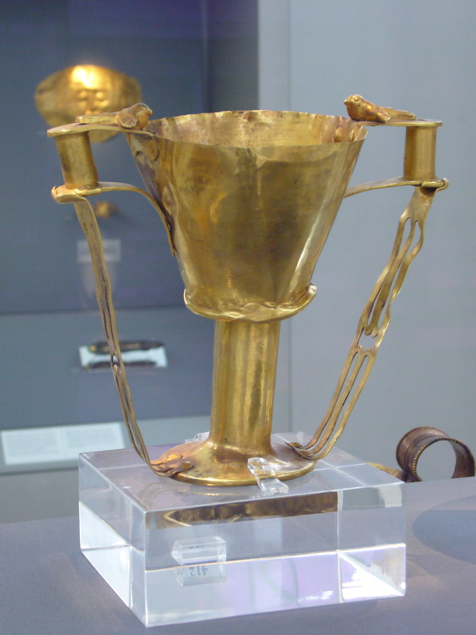 Color photograph of golden goblet with handles. There is a small bird perched on each handle.