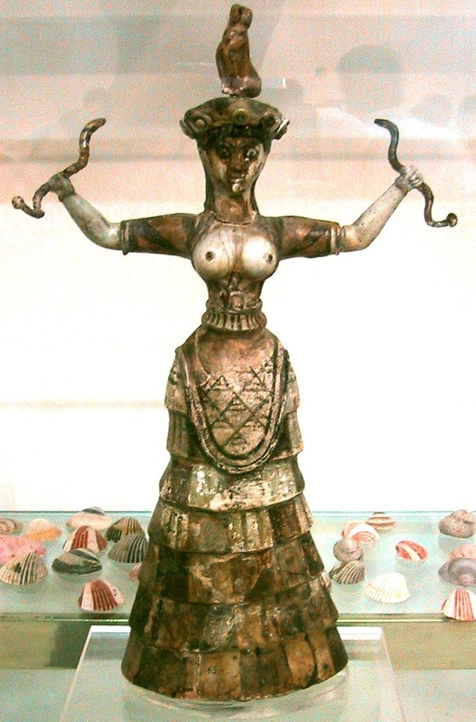 This is a color photograph of the Snake Goddess figurine. It depicts a woman with open arms who holds a snake in each hand, with a feline sitting on her head. She is dressed in a common Minoan style of clothing, a full skirt and a tunic opened at the chest to reveal her breasts.