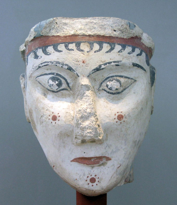 This is a color photograph of a stylized sculpture depicting a female head of a priestess, goddess, or sphinx. Her face is decorated with flower symbols and she wears an abstract headpiece.