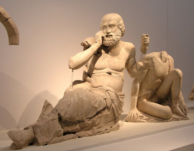 This is a photo of the seer from the east pediment of the Temple of Zeus. The seer is shirtless and wears a draped skirt-like garment. He has a beard and wears a terrified expression on his face.