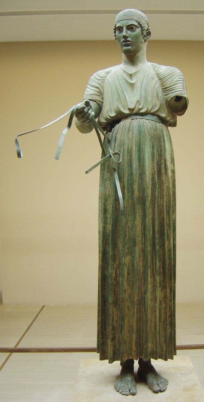 This is a photo of the bronze statue the Charioteer of Delphi, a free-standing charioteer wearing a dress and holding what appears to be the reigns that were attached to the horses that were originally part of the statue.