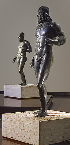 This is a photo of the Riace Warriors. Warrior A is on the right and Warrior B on the left. They are made of bronze and appear nude with idealized bodies, including prominent chiseled abdominal muscles. Both wear helmets.
