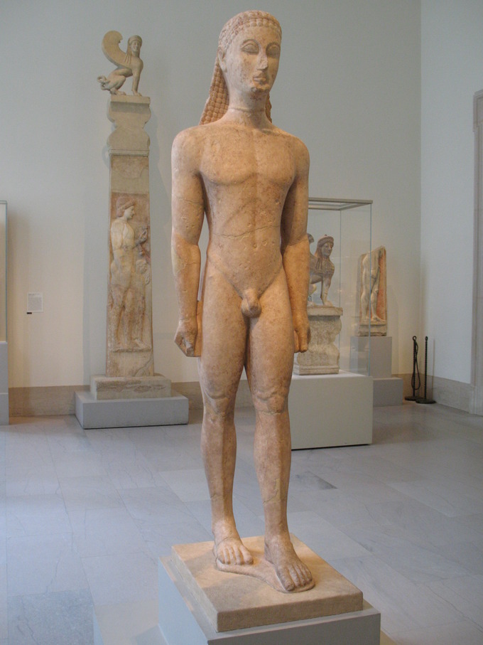 Color photograph of a marble statue of nude male Greek youth. The youth stands straight with his hands at his side and either wears a headpiece or has stylized hair.