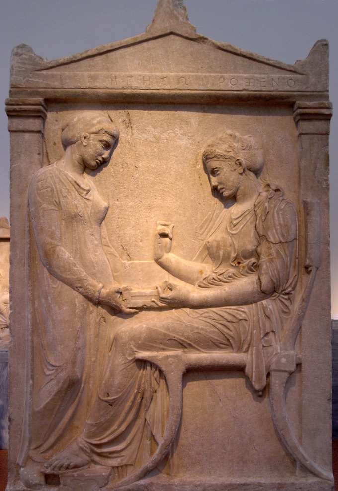 Esta es una foto de la Estela Grave de Hegeso, que representa a dos mujeres con prendas drapeadas. Una mujer está sentada y la otra se para ante ella, colocando un objeto cuadrado en su regazo.