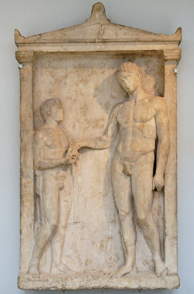 This is a photo of the Grave Stele of an Athlete, which depicts a young nude attendant and a much taller nude athlete, cocking his head and reaching for something that the attendant is holding.