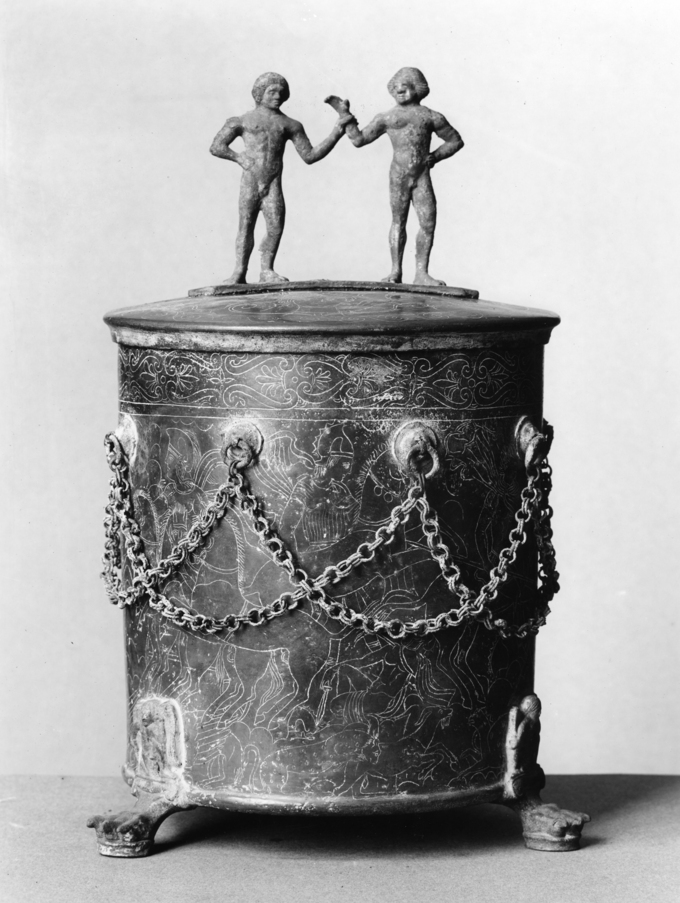 This is a photo of a cista ( a bronze circular container with a lid used to store goods). The handles are nude male statuettes.The cista is engraved with battle scenes between the Etruscans and the Gauls and decorated with a criss-crossed chains.