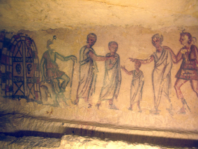 This is a photo of a fresco depicting Charun and Vanth outside the gates of the underworld. Charun is depicted as a seated male figure clothed in red. Vanth is depicted as a woman in a short tunic leads a group of deceased people dressed in white robes toward Charun and the gate.