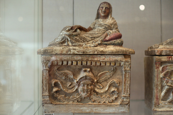 This is a photo of a terra cotta cinerary urn. A depiction of the deceased, an woman, lies across the lid wearing draped clothing that covers her head like a hood. On the bottom is a relief of her face.