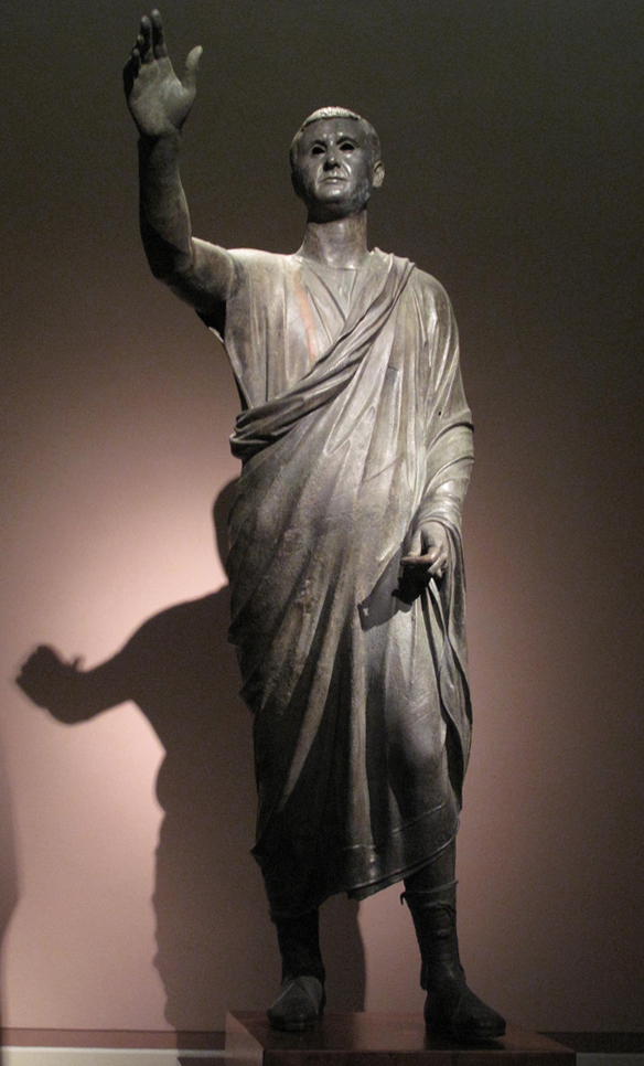 This is a photo of that statue called Aule Metele. It depicts a man wearing a short Roman toga and footwear. His right arm is raised to indicate that he is an orator addressing the public.