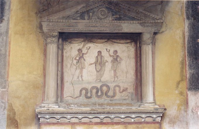 This photo shows a fresco and stucco lararium from the House of the Vettii. Two Lares (guardian deities), each holding a rhyton (cone-shaped drinking container), flank an ancestor-genius holding a libation bowl and incense box, his head covered as if for sacrifice. The snake, associated with the land's fertility and thus prosperity, approaches a low, laden altar.