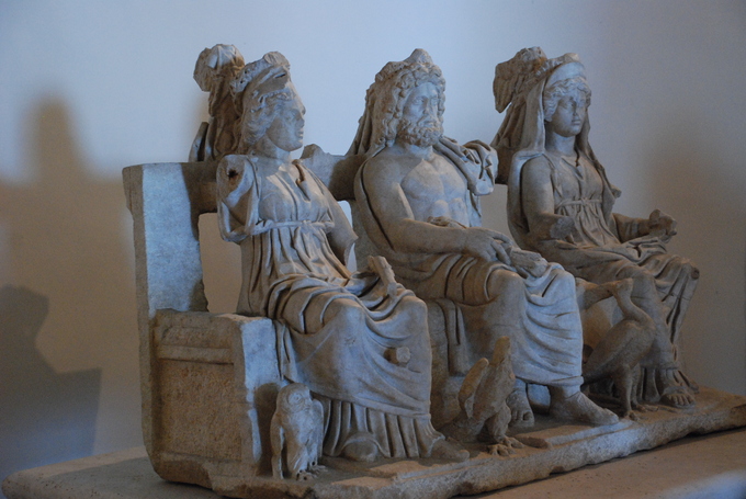 This photo is taken in the Sanctuary of Fortuna Primigenia, in Palestrina, Italy. It shows Juno, Jupiter, and Minerva seated on a bench. Juno and Minerva wear togas and have stylized hair. Jupiter is bare-chested and bearded.