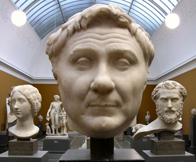 This photo shows a marble bust of Pompey the Great in a museum with other busts in the background. The bust depicts a round face and small lidded eyes. His hair is brushed back high from the forehead.