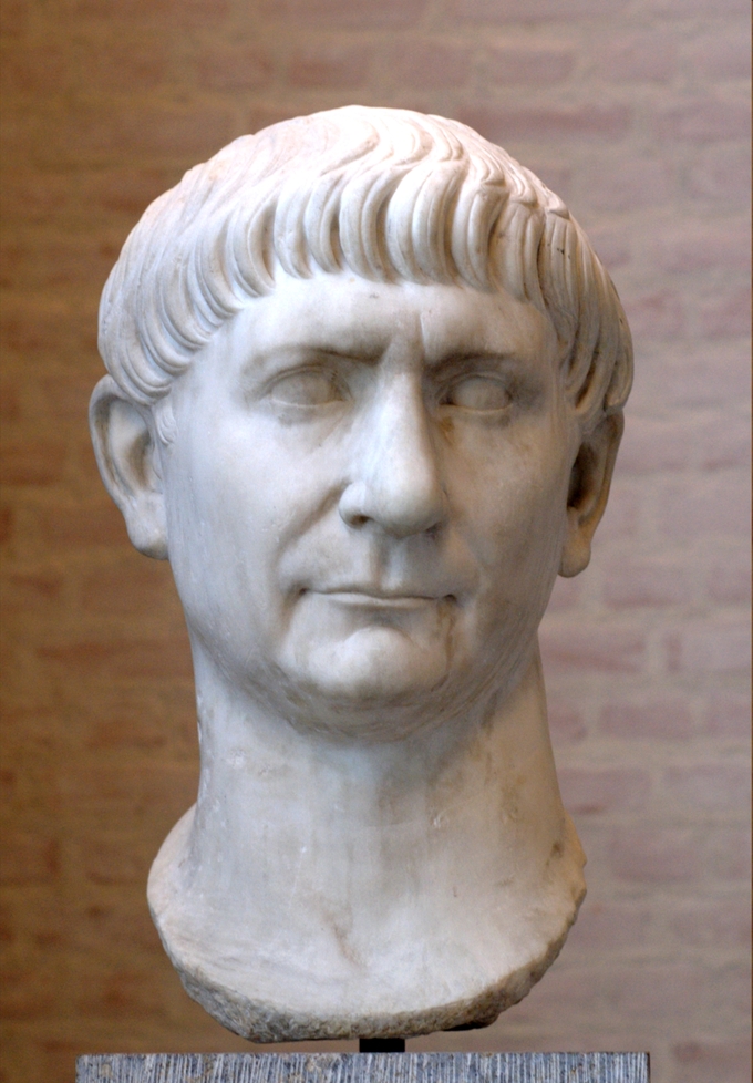 This photo shows a bust of Trajan. He has a full head of hair with bangs that stop above his eyebrows, triangular nose, and thin straight lips.