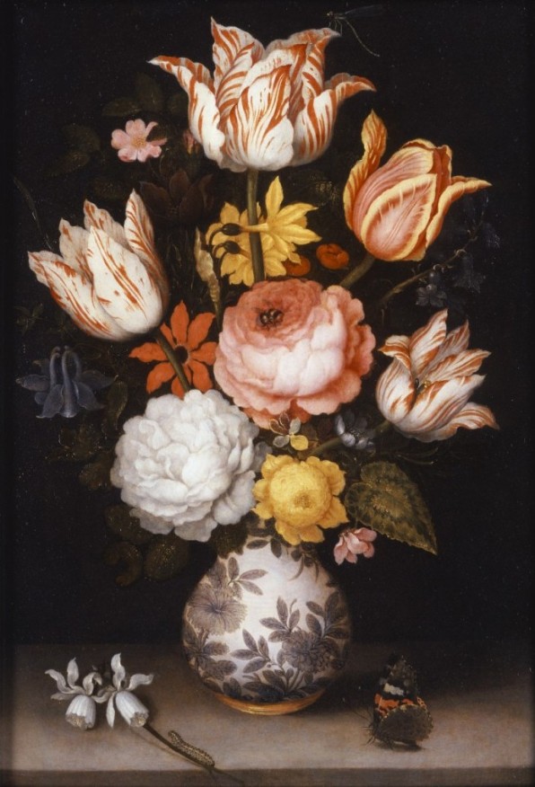 A variety of flowers in different colors are in a vase decorated with a floral design. One flower has fallen on the table, and a butterfly sits on the other side of the table.