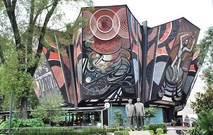 Picture of the exterior of the building. A statue of two men is in front of the building, and a colorful mural with several images covers the entire outside wall.