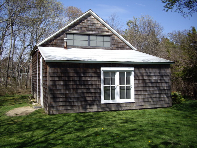 A photo of the exterior of the Pollock Barn. It is a plain, small house with dark shingles and white windows.