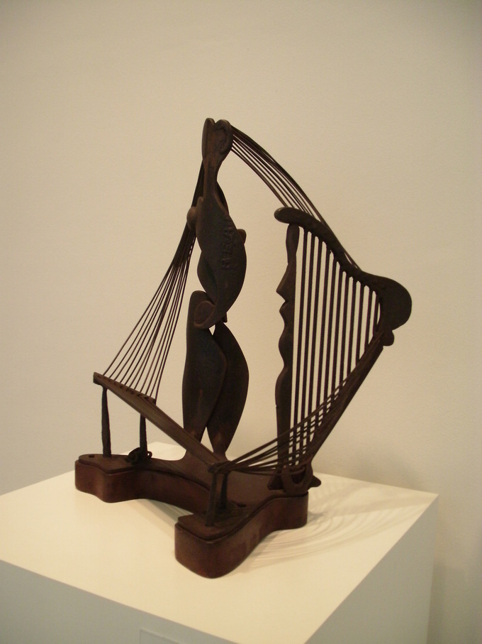 A wooden looking sculpture made up of abstract images. There is a central piece with string-like objects on either side.