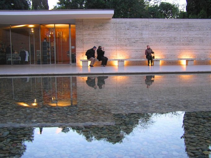 An image of the exterior of the Barcelona Pavilion. Three people sit on a bench outside. The exterior wall is simple and there are glass doors leading inside. In the foreground is a clear pool over many rocks.