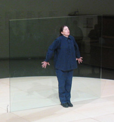 A photo of Marina Abramovic standing in front of a glass pane as part of a performance art.