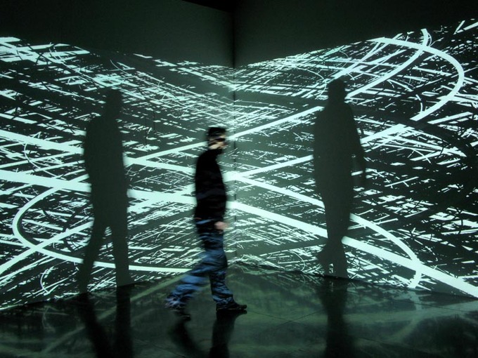 Photo of a man walking through the art installation. Green, swirly lines are projected on the wall behind him.